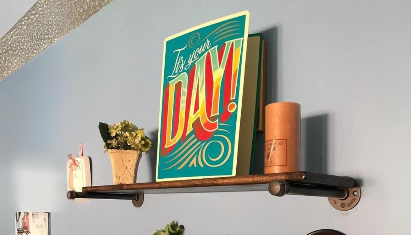 A decorative shelf with a colorful sign saying 'It's your DAY!', a small flower pot, and a candle. 