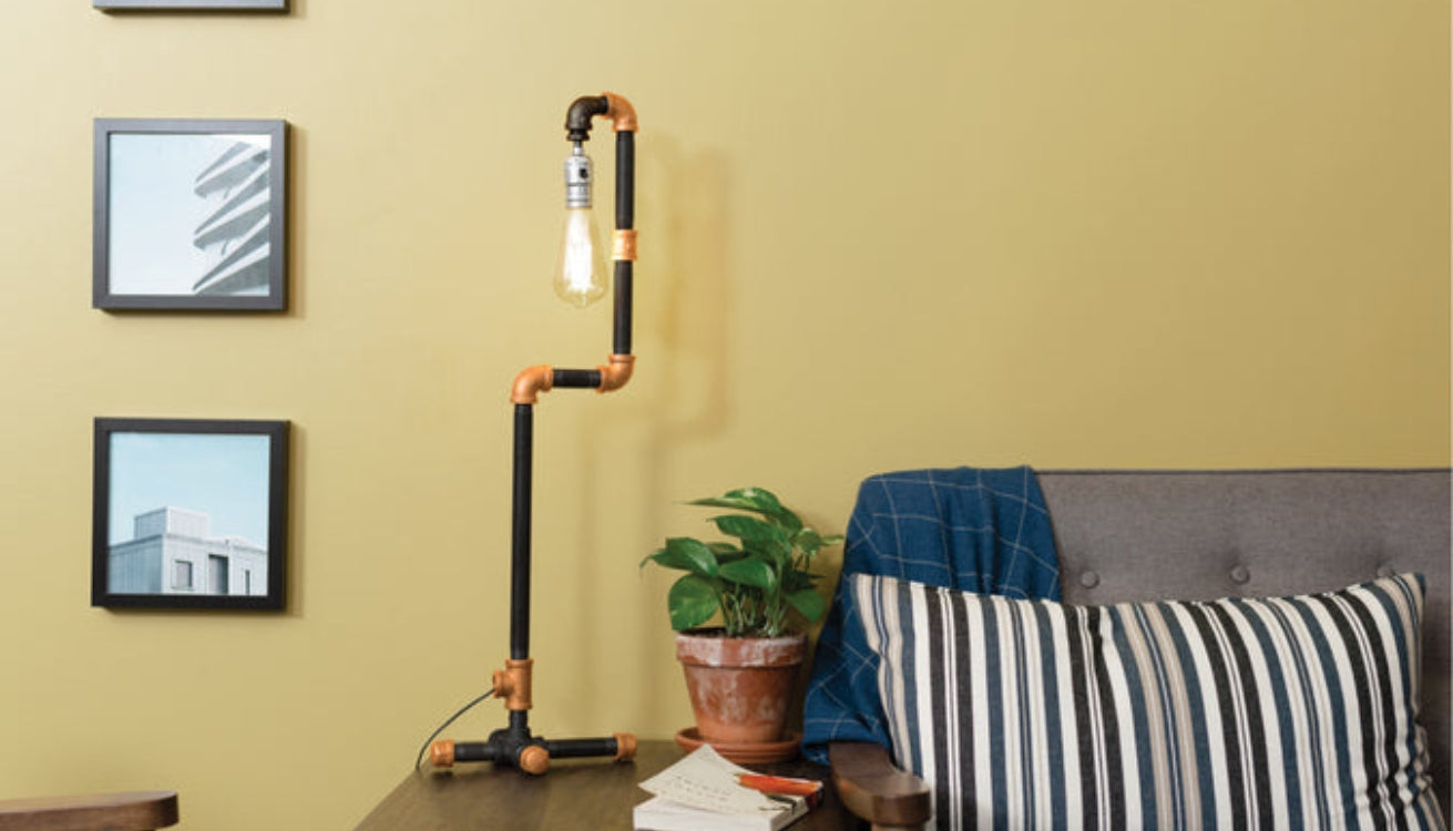 Industrial-style lamp made from matte black and brass pipes, placed on a wooden table next to a gray armchair with a striped cushion and a potted plant.