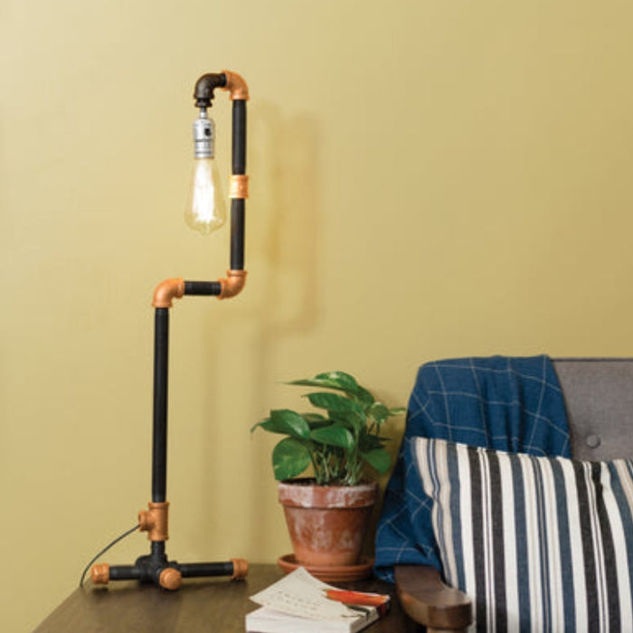 Industrial-style lamp made from matte black and brass pipes, placed on a wooden table next to a gray armchair with a striped cushion and a potted plant.