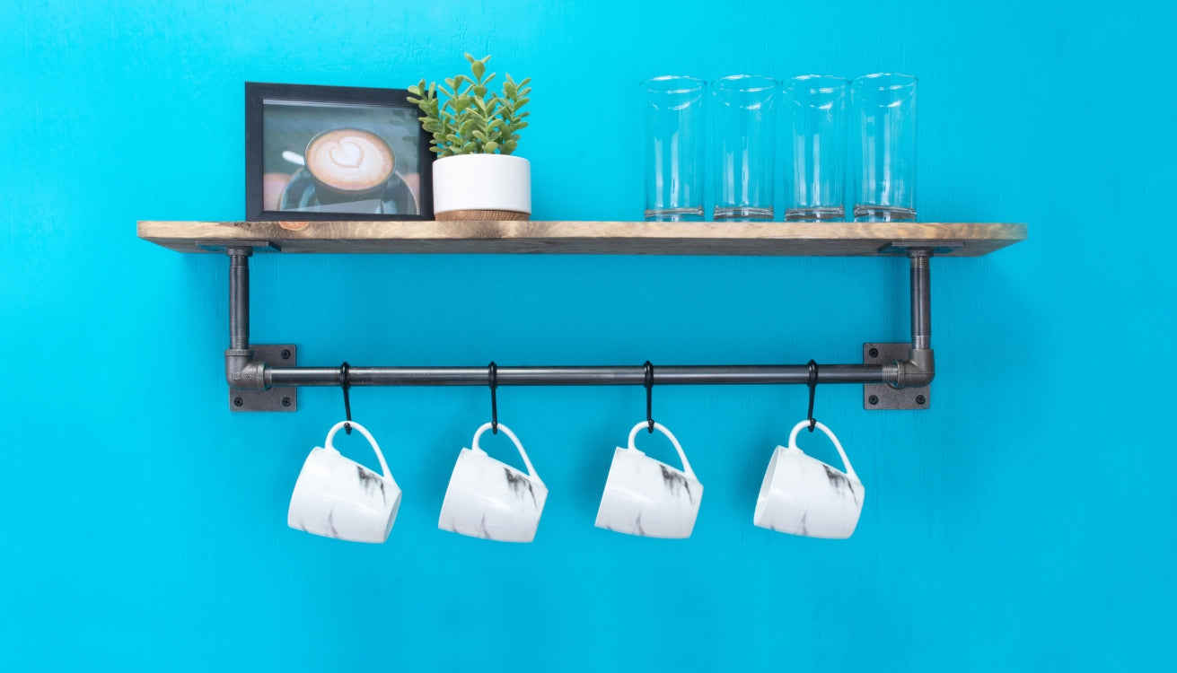 A rustic industrial pipe shelf against a vibrant blue wall, holding glasses, a framed picture, and mugs hanging from a pipe below.