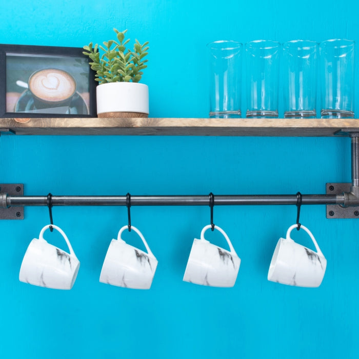 A rustic industrial pipe shelf against a vibrant blue wall, holding glasses, a framed picture, and mugs hanging from a pipe below.