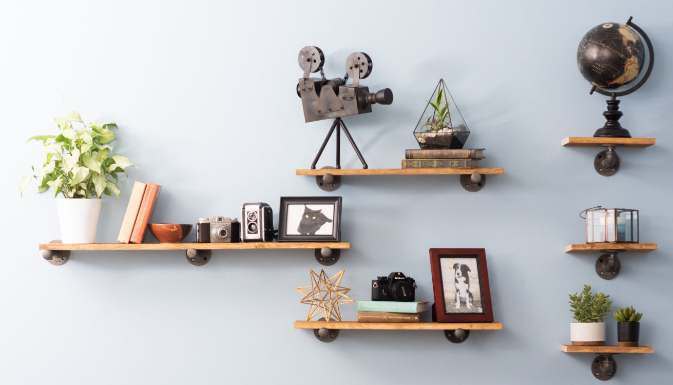 Image of a stylish floating shelf by PIPE DECOR®, displaying a globe, books, camera, and succulents against a soft green wall