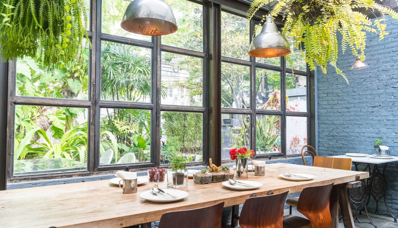 Bright coffee shop with a large window, green plants, and industrial pipe furniture. Embrace the coffee shop aesthetic with stylish pipe furniture.