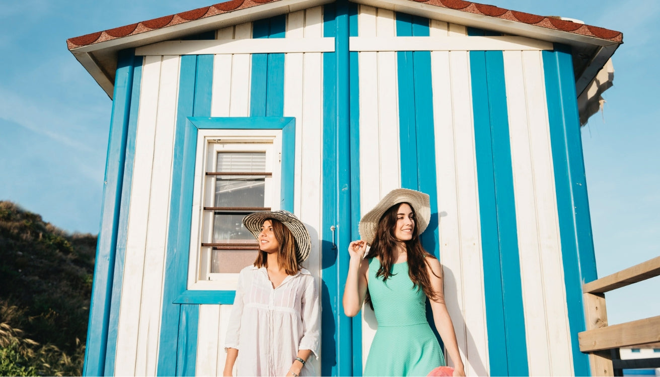 Two friends share a laugh by a vibrant blue and white striped she shed, perfect for summer vibes! Image by Freepik.