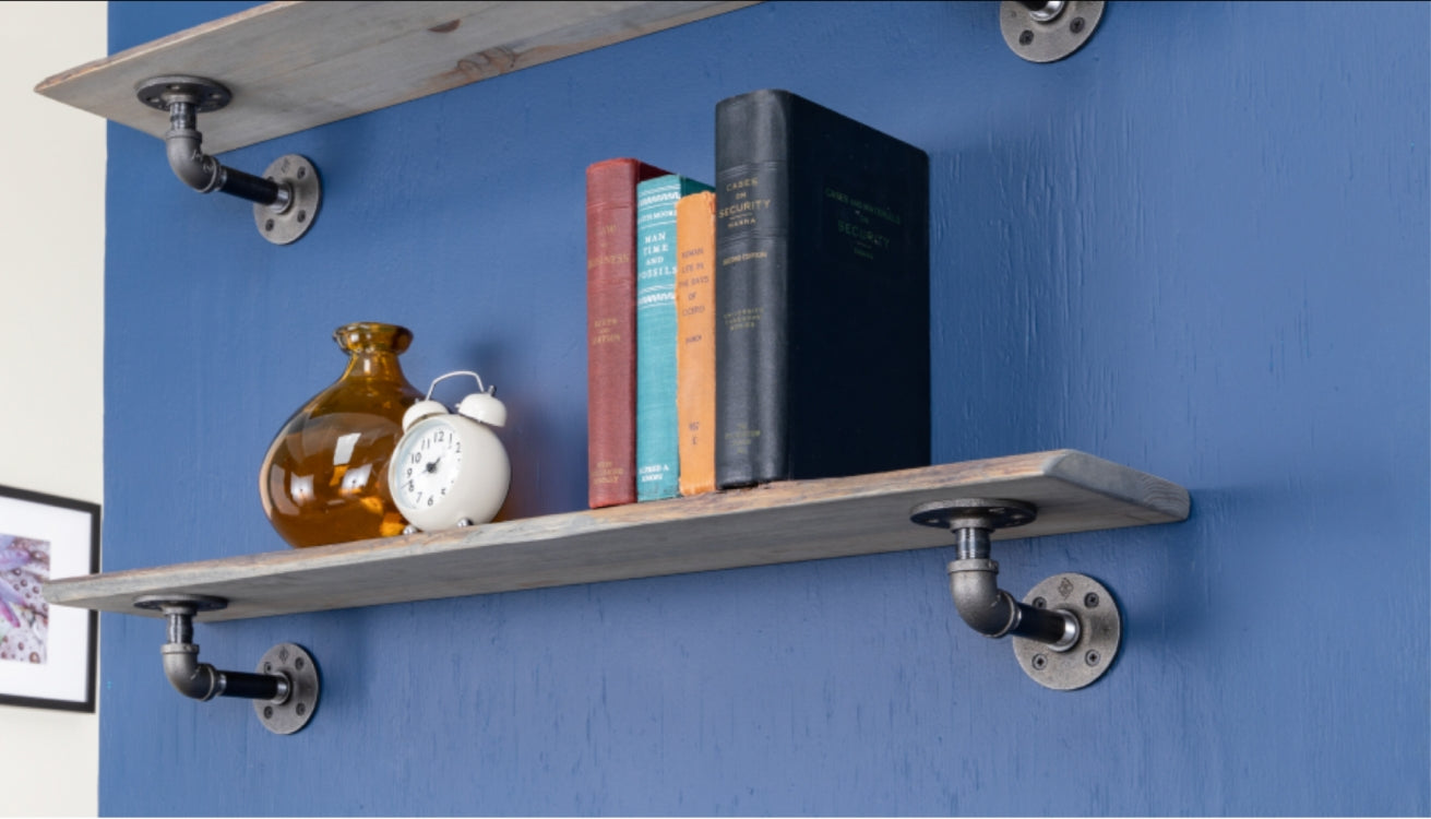 Industrial pipe shelf mounted on a blue wall, styled with books and decor items including a vintage clock and a glass vase.