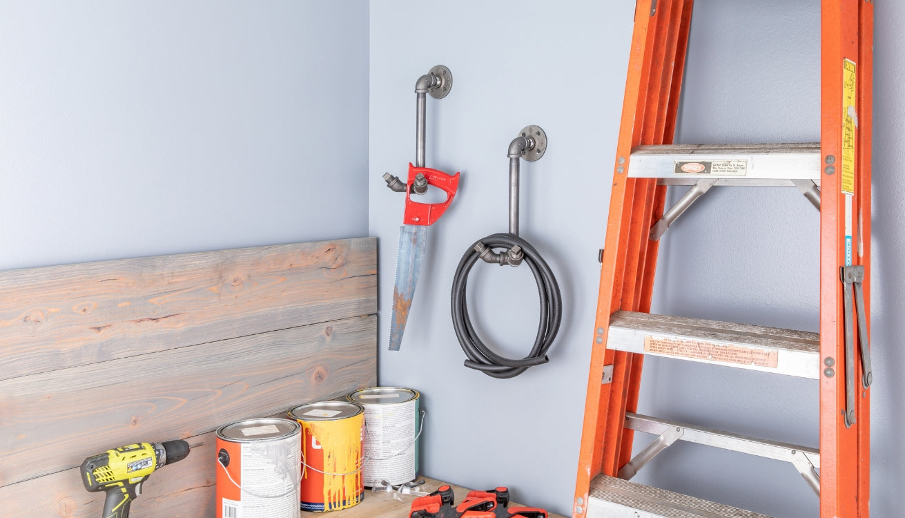 A workshop corner with pipe wall hooks holding tools and cables, an orange ladder, wood panels, and paint cans neatly arranged on the floor.